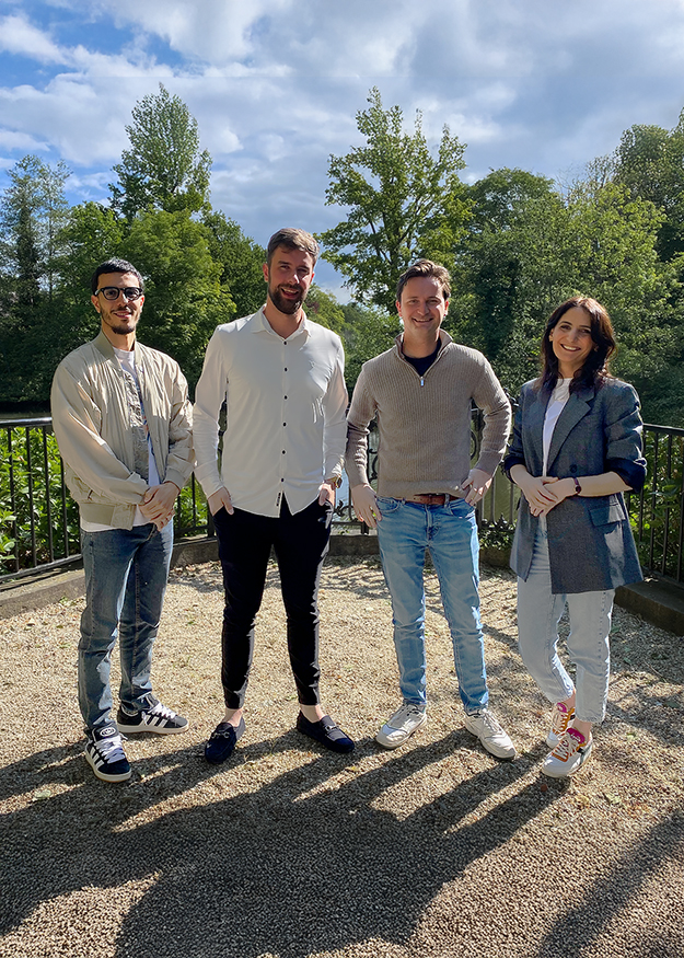 Vier collega's van ZorgDomein staan buiten in de zon, lachend en ontspannen, met een groene, bosrijke achtergrond.